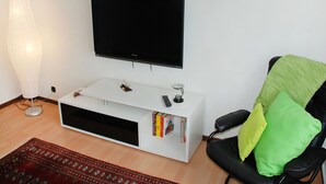 A living room with a flat-screen TV mounted on a white cabinet, a black office chair with green and yellow cushions, a patterned rug, and a floor lamp.