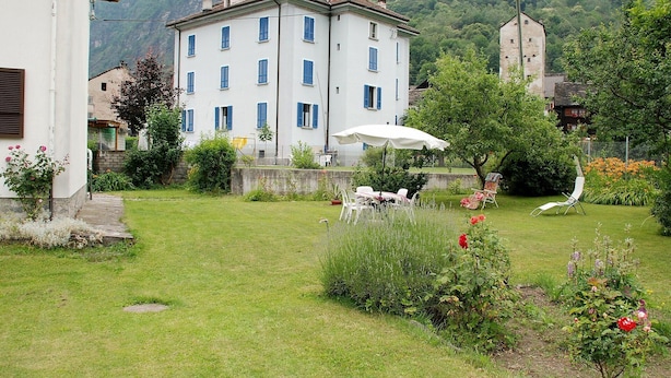 A white building with blue shutters, a garden with a table and chairs, and a mountain backdrop.