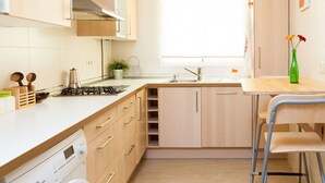 A compact kitchen with a gas stove, a sink, and wooden cabinets. There is a window providing natural light and a small table with a vase of flowers.