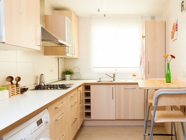 Une cuisine compacte équipée d’une cuisinière à gaz, d’un évier et de meubles en bois. Une fenêtre laisse entrer la lumière naturelle et une petite table est agrémentée d’un vase de fleurs.