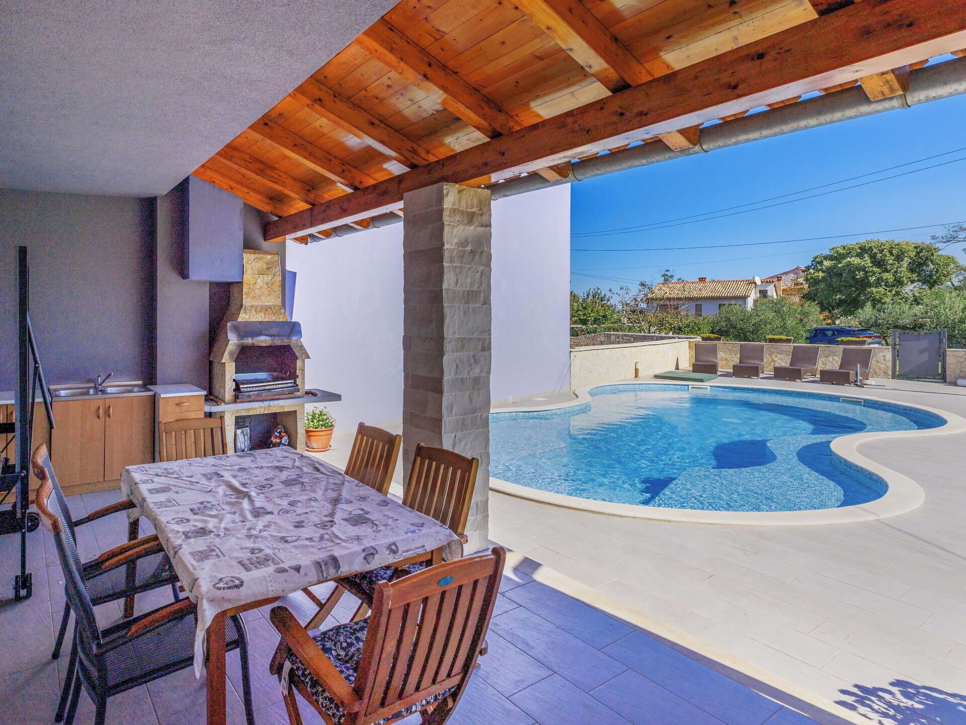 An outdoor patio with a pool, wooden furniture, a grill, and a view of a house in the background.