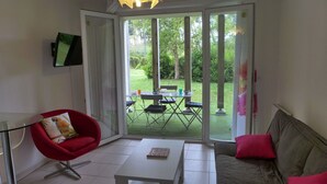 A living room with a red chair, a grey sofa, a white table, and a large sliding glass door leading to an outdoor area with a table and chairs. - Lovely apartment for 4 people with pool, WIFI, TV and terrace (Ondres)
