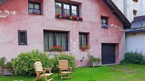 Un bâtiment rose orné de jardinières aux fenêtres, deux chaises en bois devant l’entrée et une pelouse verdoyante.
