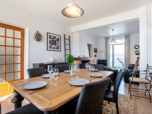 A dining area with a wooden table set for six, surrounded by chairs. There is a clock and a framed photo on the wall, and a ladder leaning against the wall.