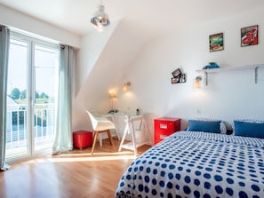 A bedroom with a bed, a desk, and a chair. There is a window with curtains, a red cabinet, and a wall with pictures.