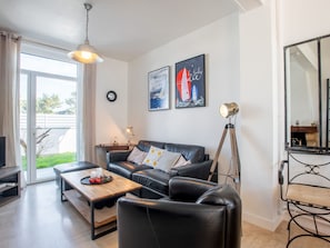 A modern living room with a black leather sofa, a wooden coffee table, and a floor lamp. There are two framed pictures on the wall, a clock, and a large mirror.