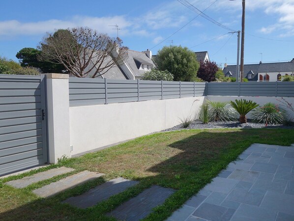 A residential area with a grey fence, a paved pathway, and a grassy area with small plants.