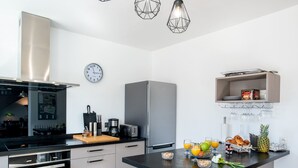Et moderne køkken med indbygget ovn, kaffemaskine og køleskab. Spiseplads med to stole og et bord dækket med frugt og glas.