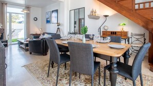 Spiseområde med et træbord dækket til seks personer, omgivet af seks stole. Et stort spejl hænger over bordet, og der er en pendellampe samt en trappe, der fører til en etage ovenover.