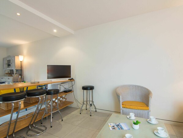 A modern kitchen with a dining area, bar stools, a television, and a wicker chair.