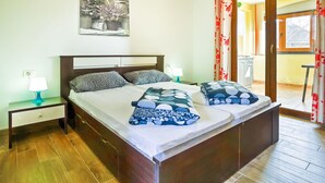 Ein Doppelbett mit gemusterten Kissen, ein Nachttisch mit Lampe, ein Wandbild und ein Blick ins Freie durch eine Tür.
