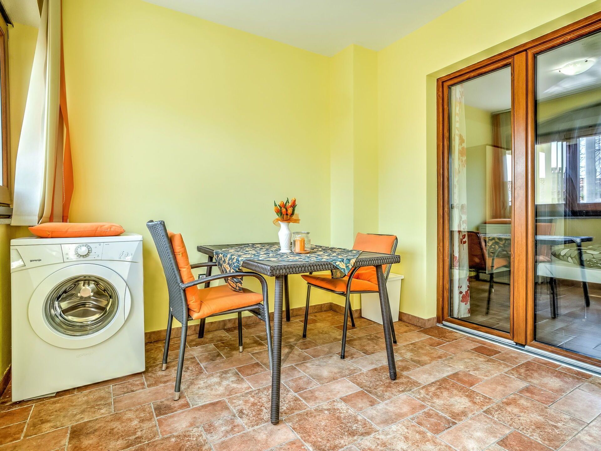 A compact living space with a washing machine, a dining table with chairs, and a sliding glass door leading to a balcony.