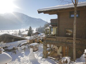Een houten chalet met een sneeuwbedekte dak, een houten terras, en een uitzicht op besneeuwde bergen op de achtergrond.