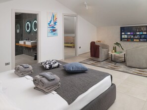 Et moderne hotelværelse med seng, badeværelse, opholdsområde og et fjernsyn, der viser en streamingtjeneste.