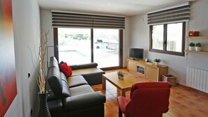 A modern living room with a sofa, a red chair, a coffee table, a TV on a wooden cabinet, and large windows with blinds. - Beautiful private villa for 8 guests with WIFI, private pool, TV, patio and pets allowed (L'ampolla)