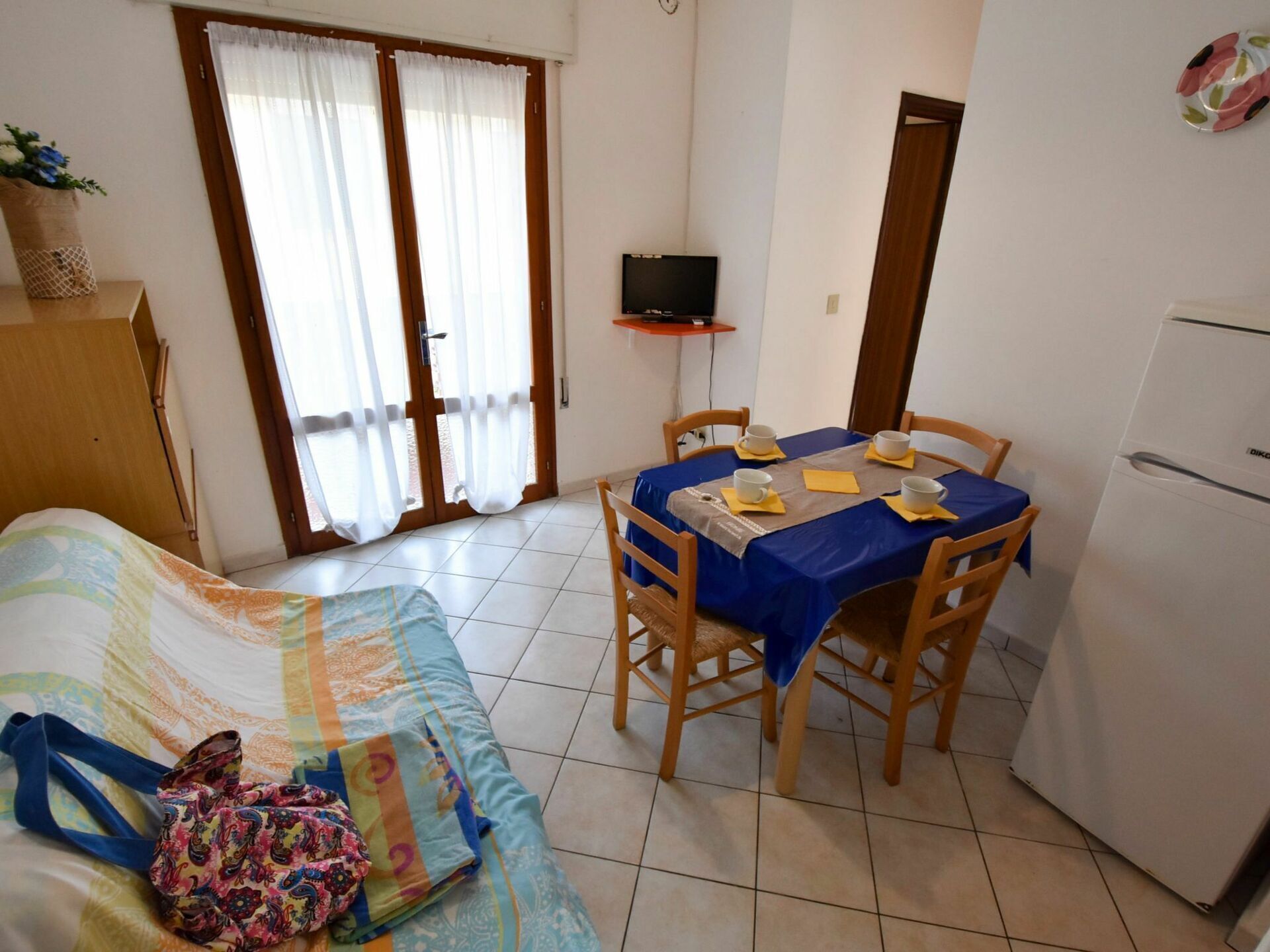 A compact living space with a bed, a dining table with chairs, a television, and a refrigerator.