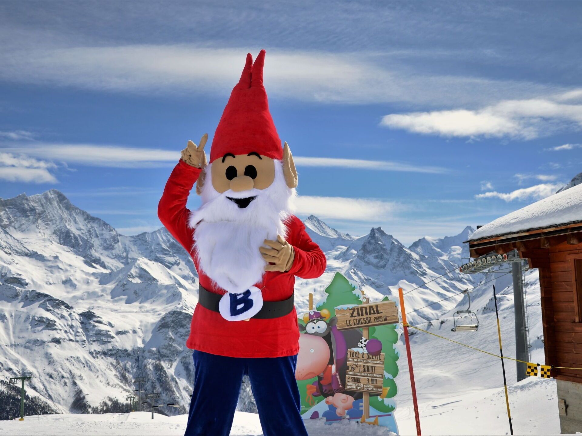 Une personne déguisée en Père Noël se tient dans une région montagneuse enneigée, avec un télésiège et un chalet en bois en arrière-plan.