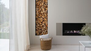 Uma sala de estar moderna com lareira, uma exibição de lenha fixada na parede, um cesto de vime e uma mesa de vidro.