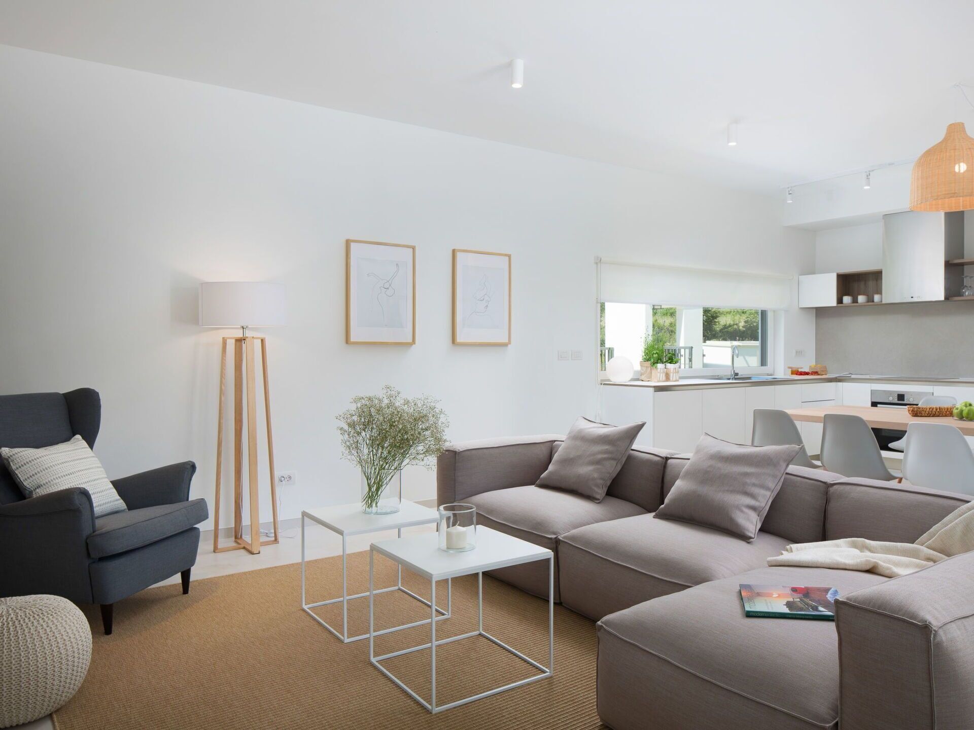 Un salon moderne avec un canapé, un fauteuil, une table basse et une lampe sur pied. On aperçoit une cuisine à l’arrière-plan, avec une fenêtre et un luminaire suspendu.