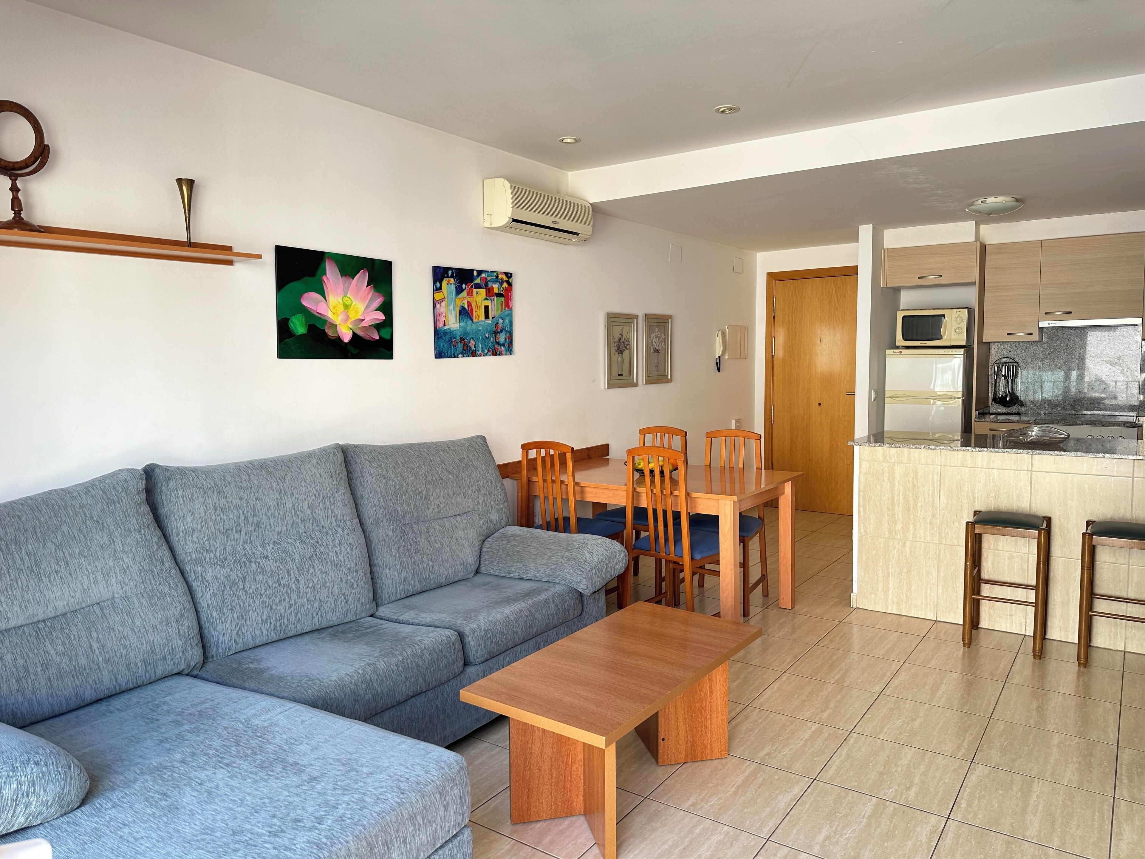 A modern living room with a grey sofa, wooden dining table, and chairs. There is a kitchen area with a microwave, stove, and cabinets.