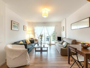 A spacious living room with a sofa, a white armchair, a dining table with a bowl of fruit, and a television. - Stunning  villa for 4 guests with WIFI, TV, terrace and pets allowed (Mimizan)