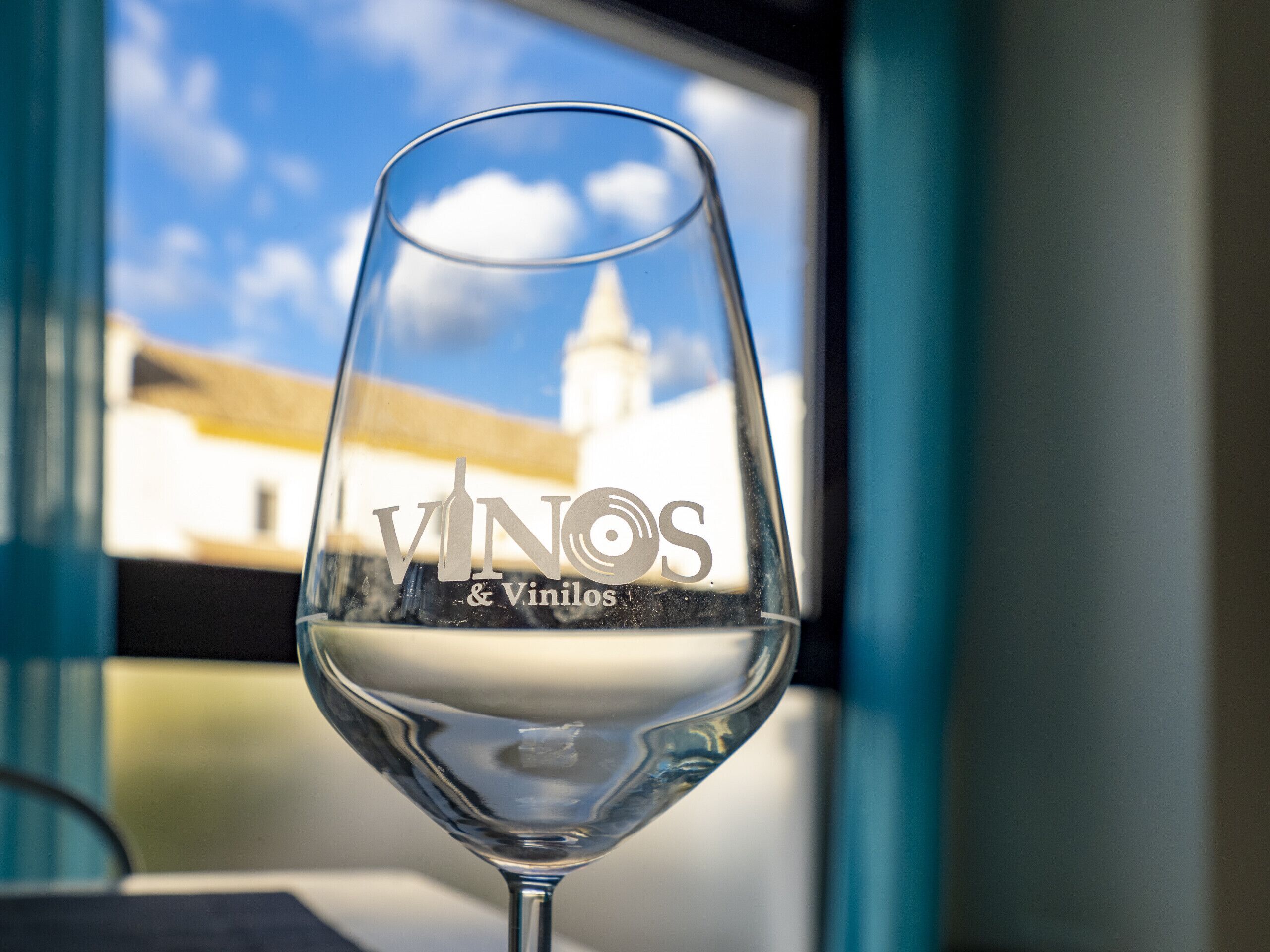 A wine glass with the inscription "VINOS & Vinilos" on it, placed in front of a window with a view of a building and a clear sky.