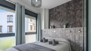 Een moderne hotelkamer met een groot bed, grijs beddengoed, een houten accentmuur en uitzicht op gebouwen door een raam met grijze gordijnen.