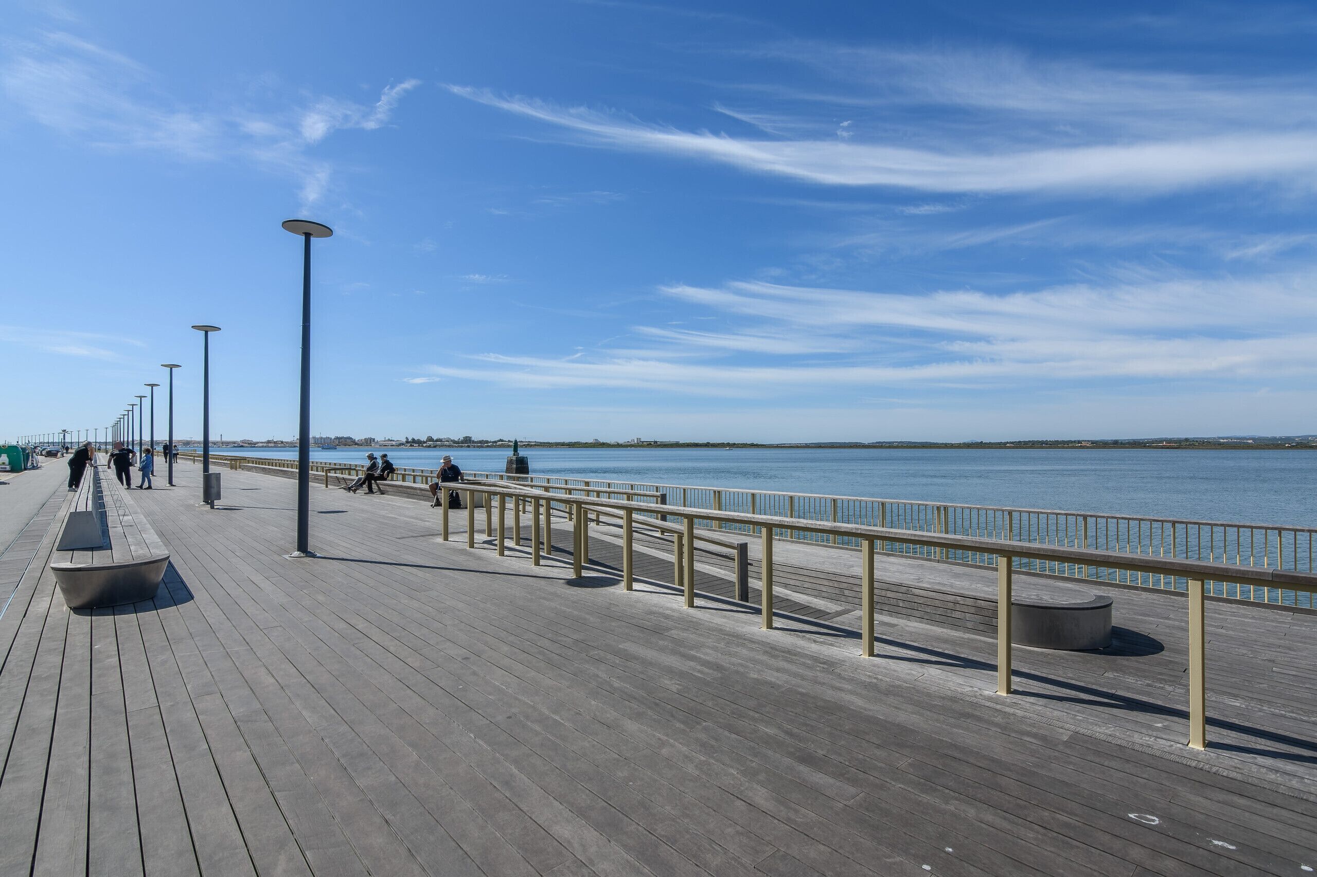 A wooden boardwalk with railings, benches, and street lamps along a waterfront.