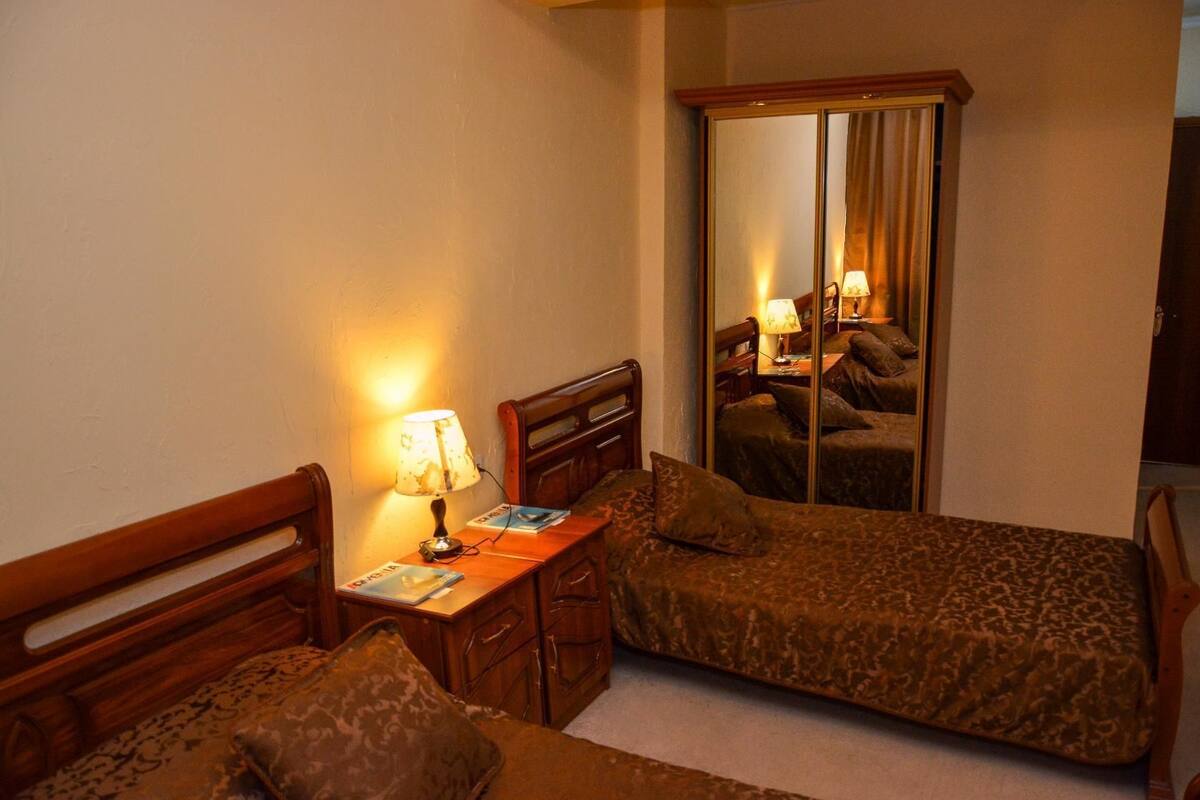 A small, cozy room with two single beds, a wooden nightstand with a lamp, a mirror, and a small table with a magazine.
