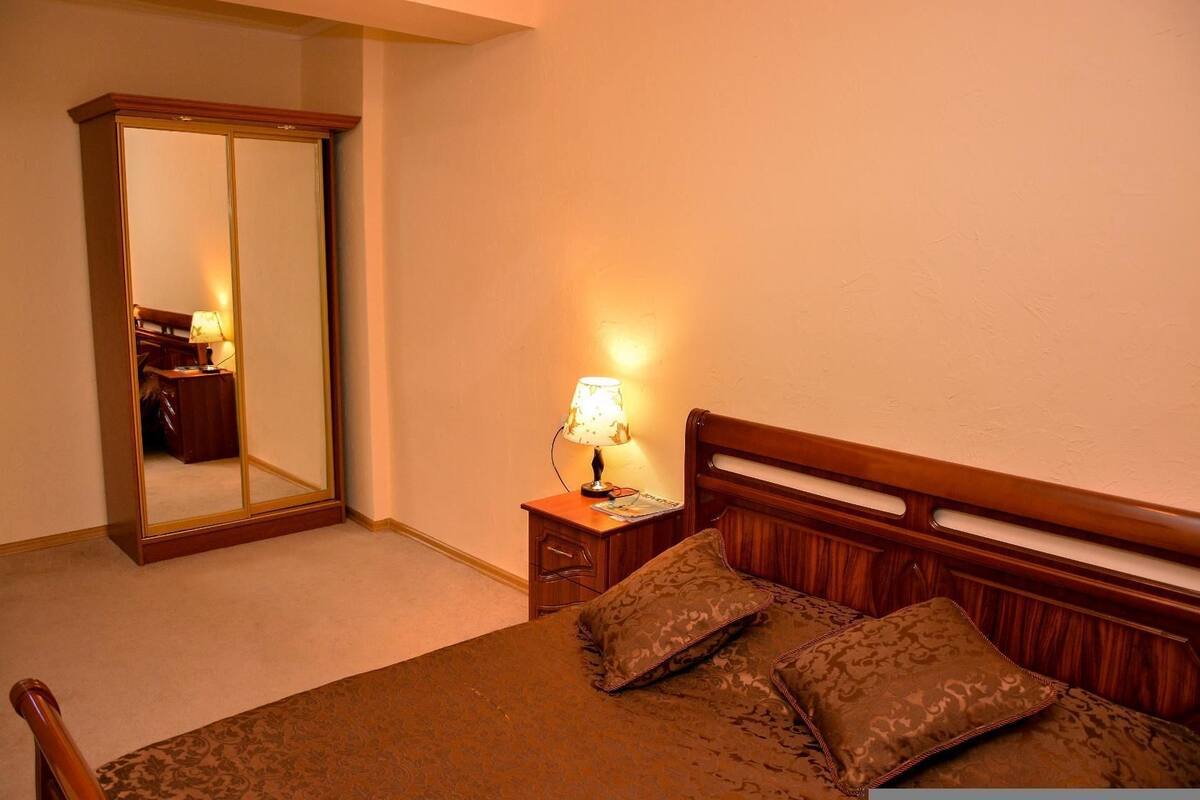 A hotel room with a bed, a nightstand with a lamp, a wooden headboard, and a full-length mirror.