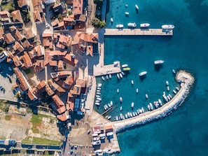 An aerial view of a coastal town with tightly packed buildings, a marina with numerous boats, and a curved breakwater extending into the sea.