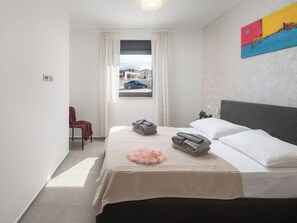 Quarto com cama, janela com cortinas, uma cadeira e um quadro na parede.