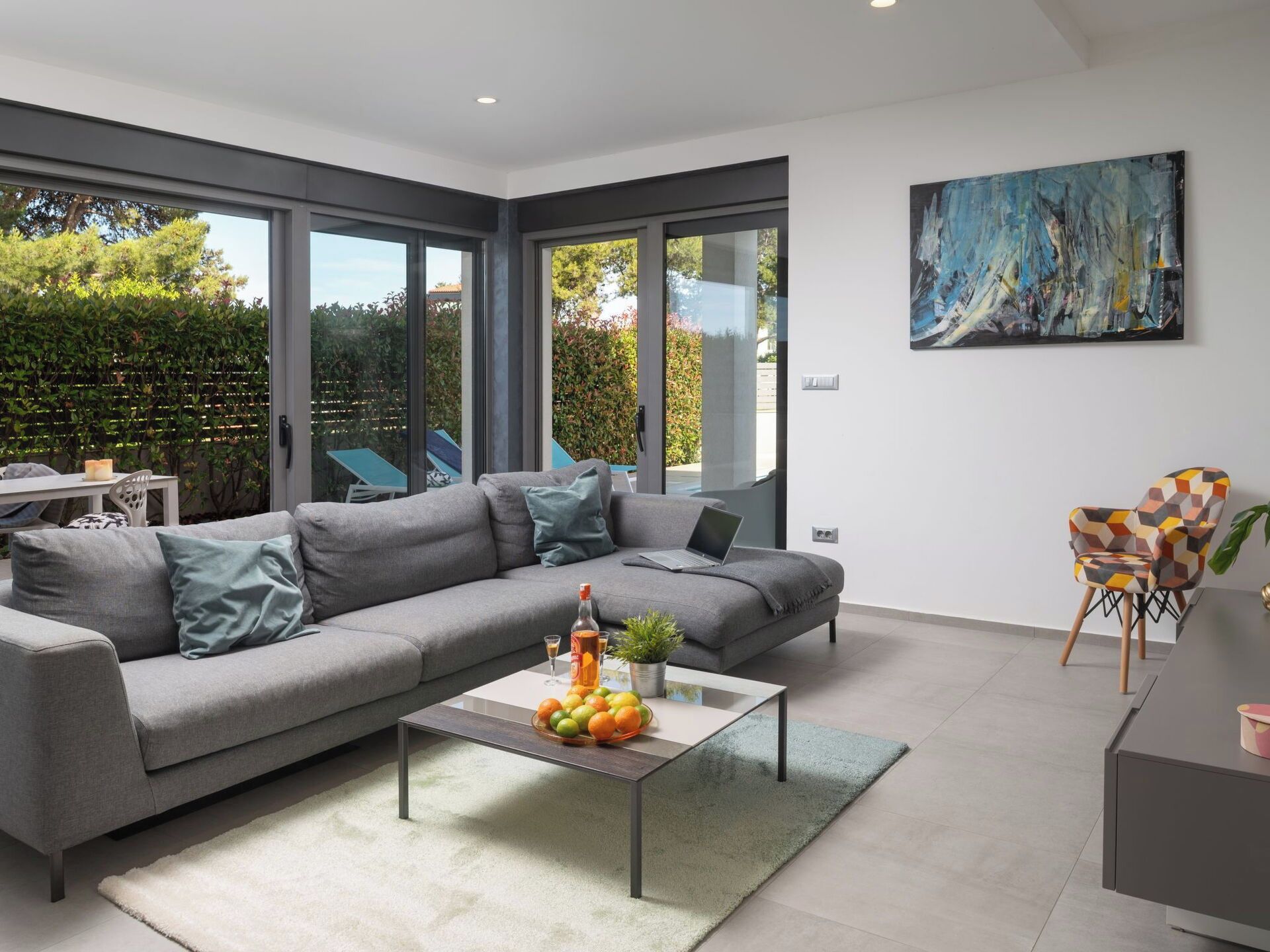 A modern living room with a grey sofa, a coffee table with fruits and a bottle, and a painting on the wall.