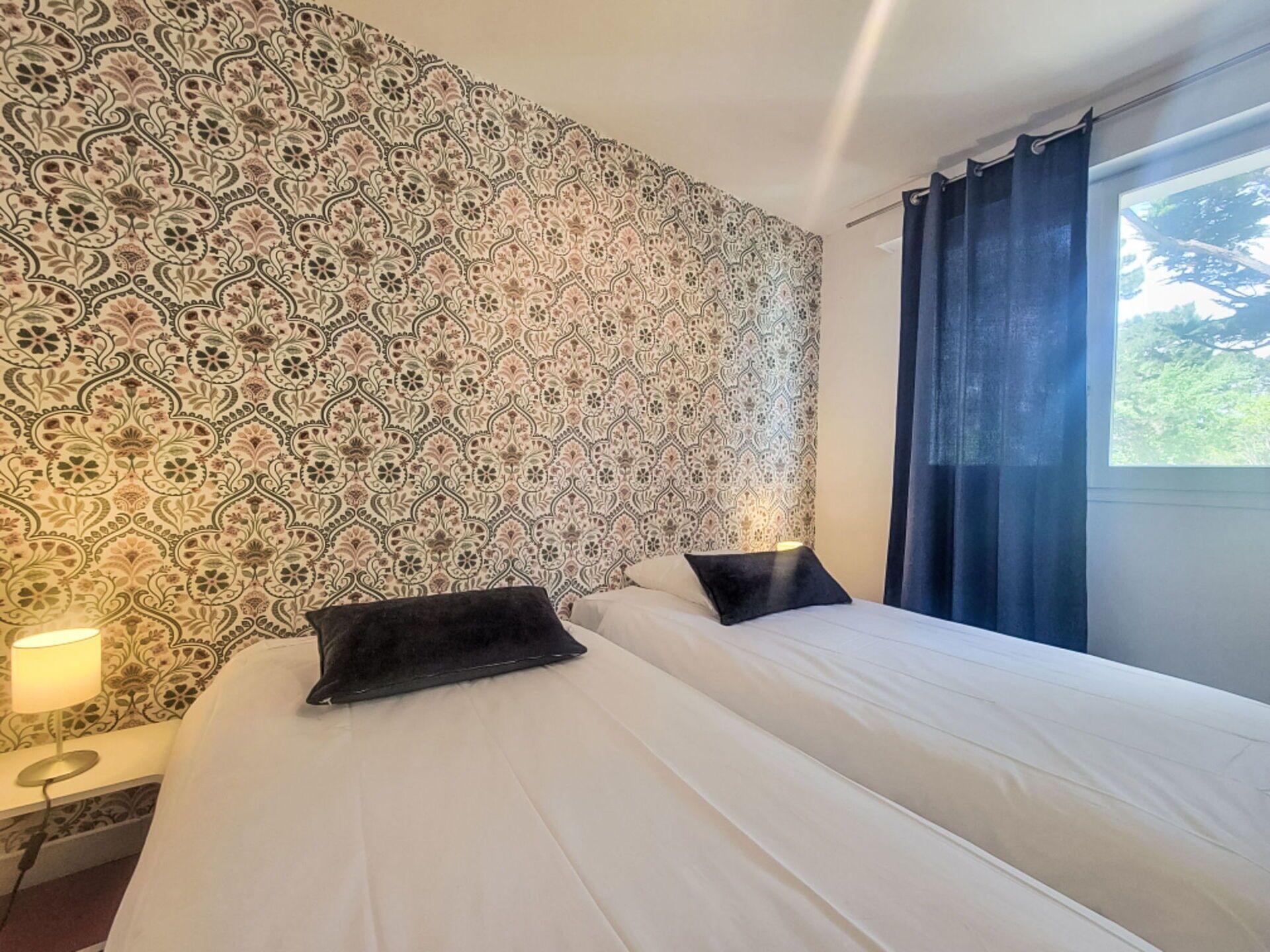 A hotel room with a patterned wallpaper, a bed with white linens, two black pillows, a bedside lamp, and a window with curtains.