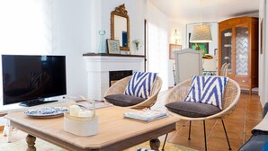 Una sala de estar con chimenea, sillas de mimbre, un televisor de pantalla plana y un comedor con mantel individual estampado.