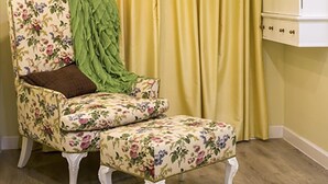 Une pièce avec un fauteuil et un pouf à motifs floraux, une armoire blanche et des rideaux jaunes.
