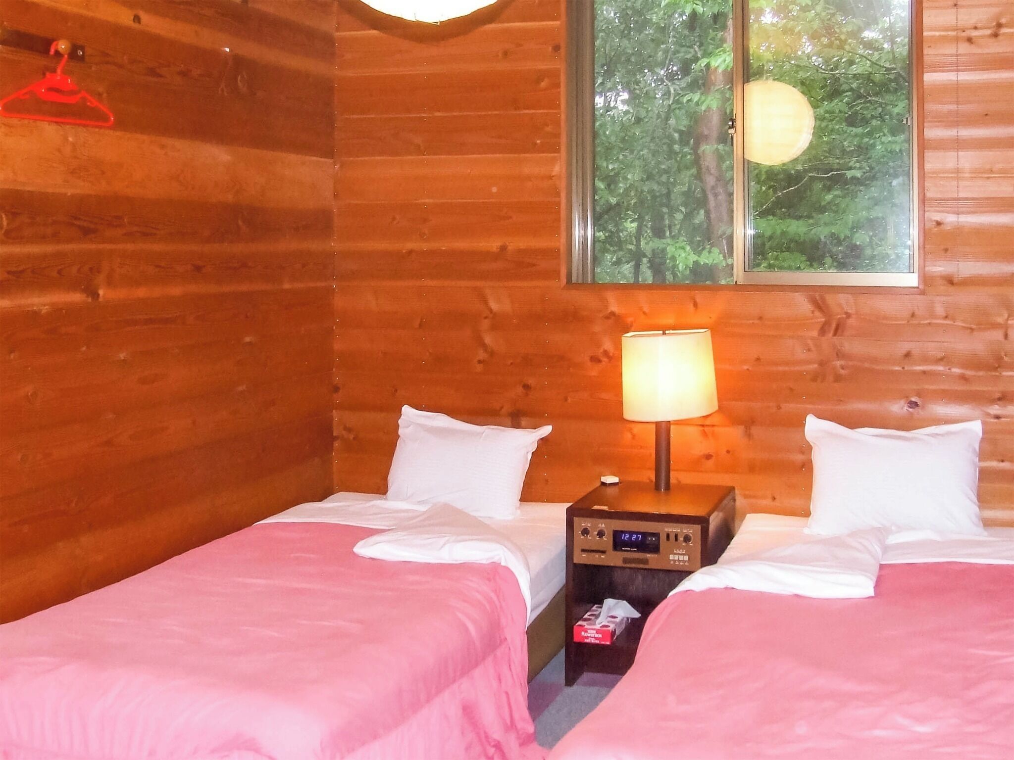Ein blockhauskaminzimmer mit zwei Betten, einem Nachttisch mit Lampe, einer Uhr und einem Fenster mit Blick auf Bäume.