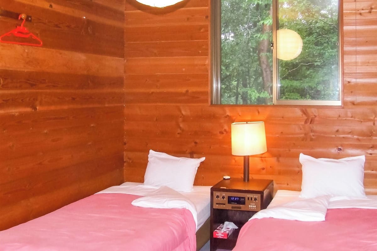 Een kamer in chaletstijl met twee bedden, een nachtkastje met lamp, een klok en een raam met uitzicht op bomen.