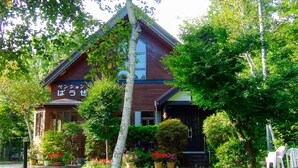 A traditional Japanese house with a sloped roof, surrounded by lush greenery and potted plants.
