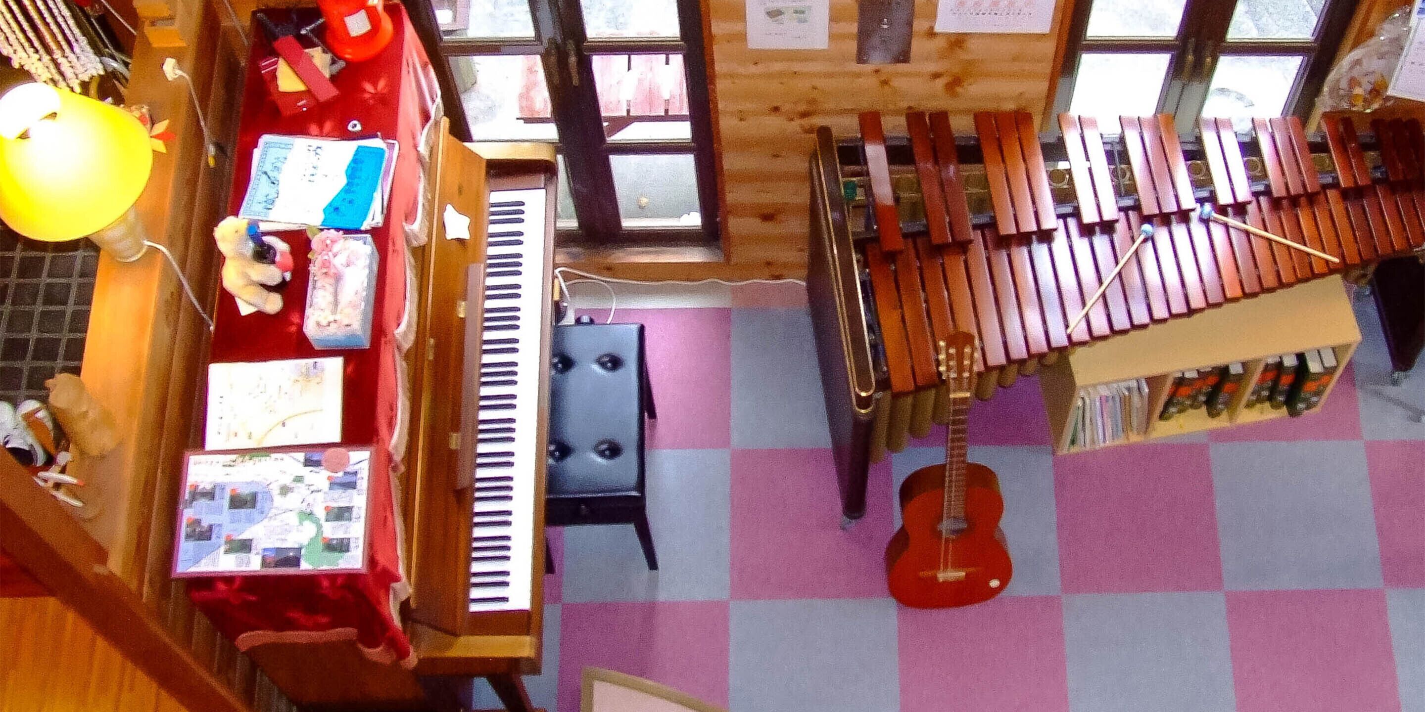 A room with a wooden floor, a red piano, a small table with a teddy bear, and a guitar hanging on the wall.