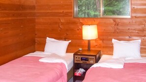 A wooden cabin room with two beds, a nightstand with a lamp, and a window with a forest view.