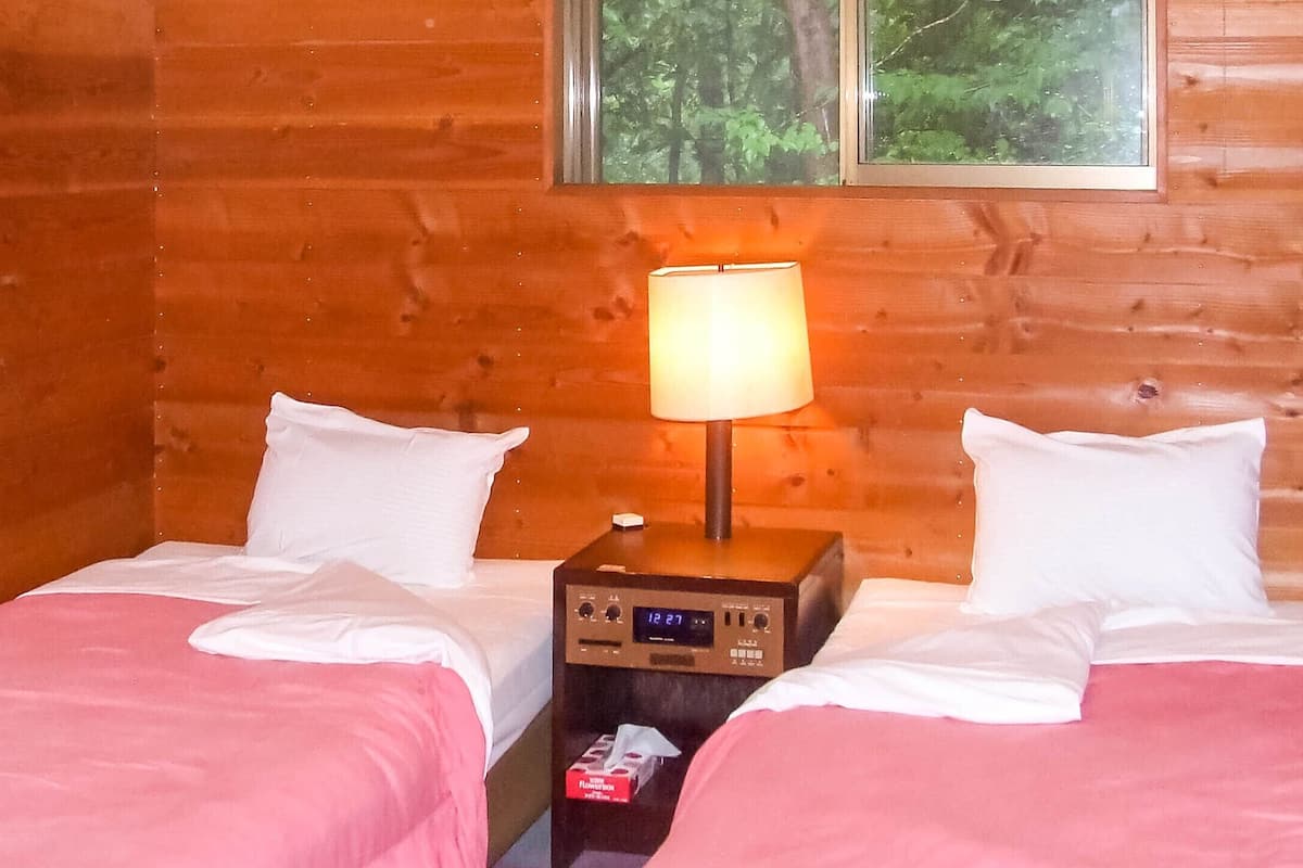 Een houten kamer met twee bedden, een nachtkastje met lamp en een raam met uitzicht op een bos.