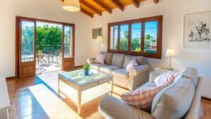 A spacious living room with a wooden ceiling, a sofa set, a glass coffee table, and a view of the outdoors through large windows.