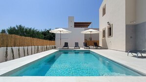 A rectangular swimming pool with clear blue water, surrounded by a tiled deck. There are four lounge chairs and two umbrellas providing shade.