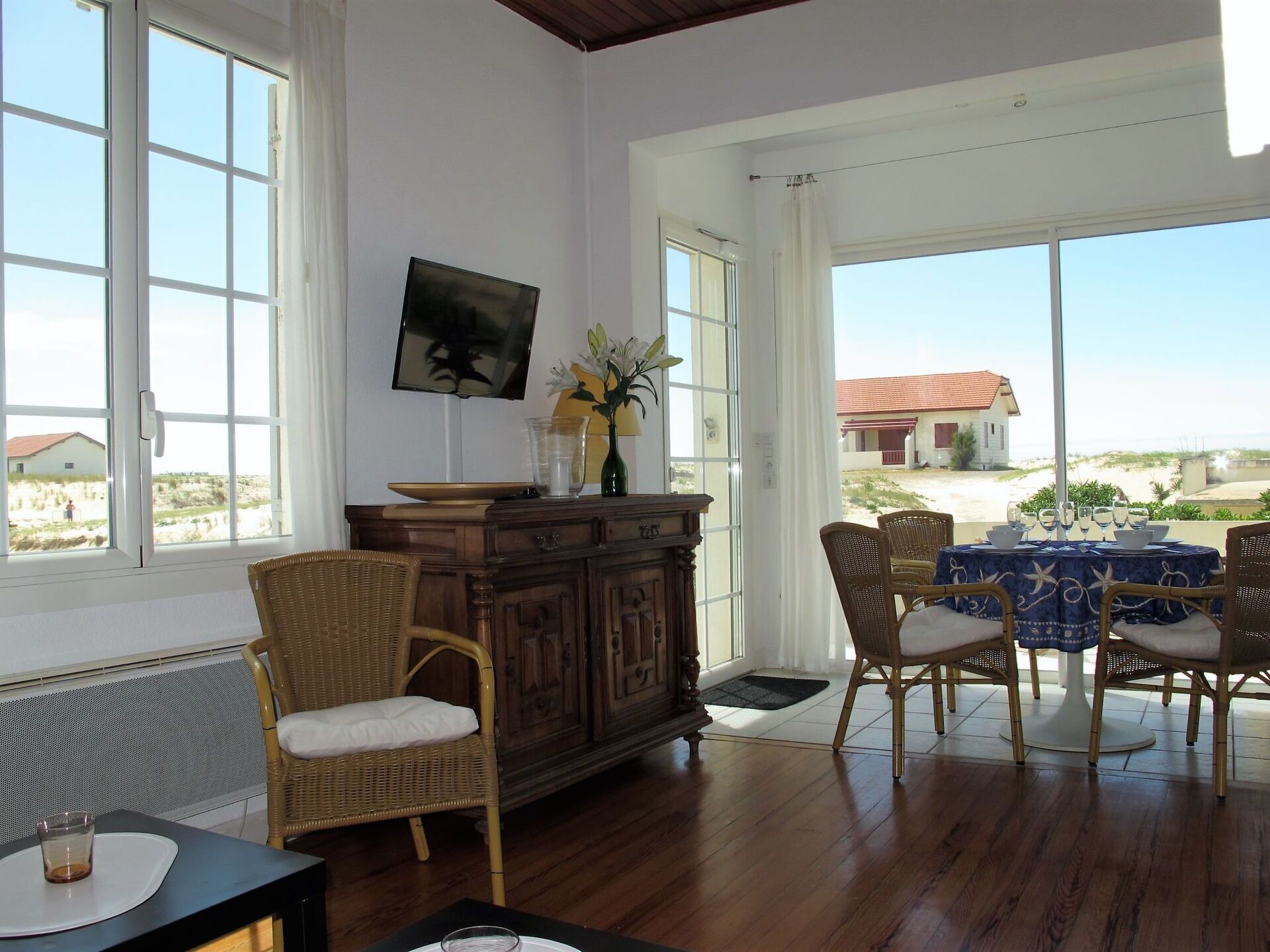 A room with a dining area, wooden furniture, a TV on the wall, and a view of the beach.