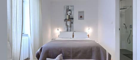 A modern bedroom with a large bed, bedside lamps, a bedside table, a woven basket, and a glass shower enclosure.