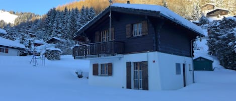 A wooden chalet with a balcony, surrounded by snow-covered trees and a mountain backdrop.