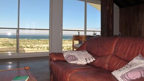 A leather sofa with cushions, a wooden coffee table with a book, and a view of the beach through large windows. - Nice apartment for 4 guests with WIFI, TV and balcony (Mimizan)