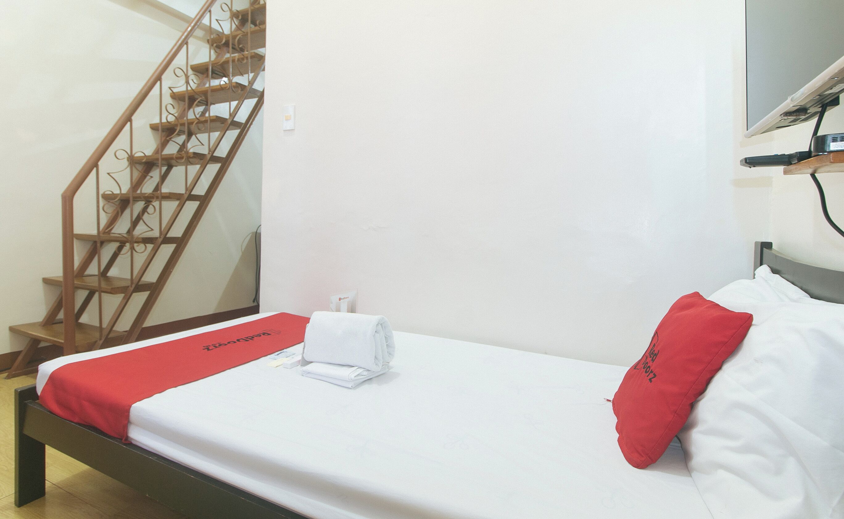 A hotel room with a bed, a red and white bedspread, a red pillow, a white towel, and a wooden staircase.