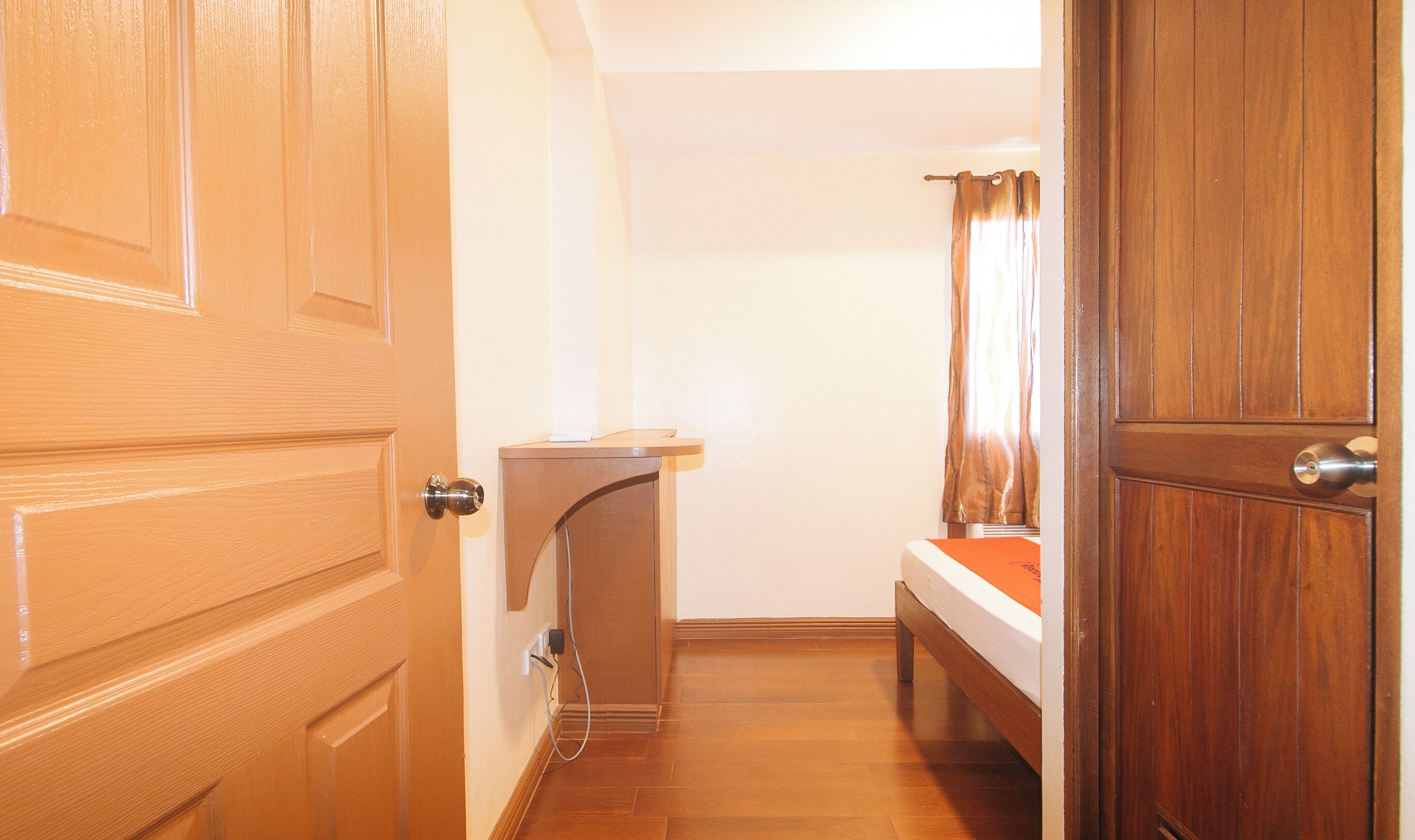 Una habitación estrecha con una puerta de madera, una cama con colcha naranja, una mesita pequeña y una cortina.